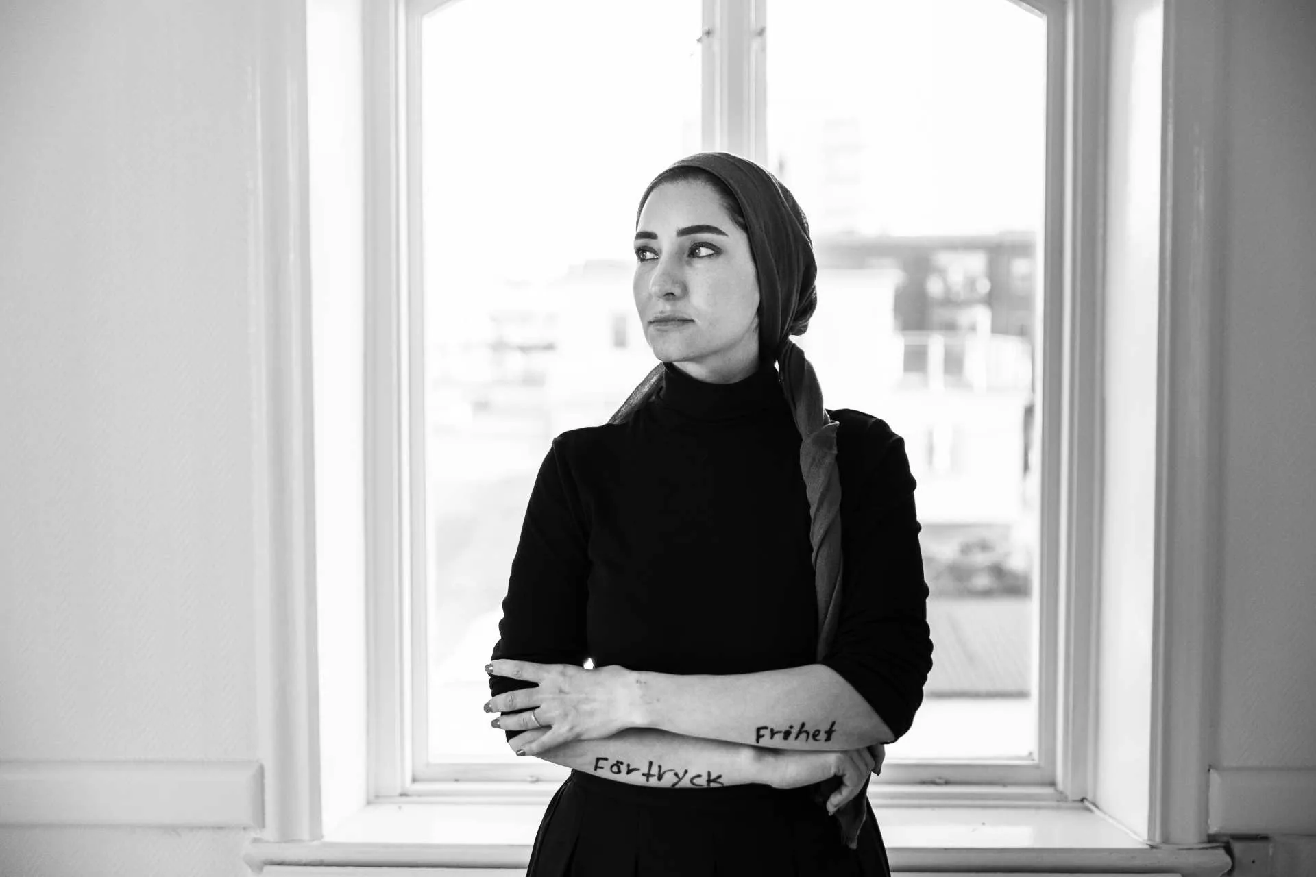 Black-and-white photograph: a woman in a headscarf and black polo neck stands against a bright window with her arms crossed; the words “Förtryck” and “Frihet” are written by hand along her forearms.