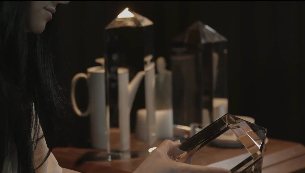 Film still: a woman in profile holds a faceted glass form beside a table of glass tower shapes, a jug and a cup, lit against a dark background.